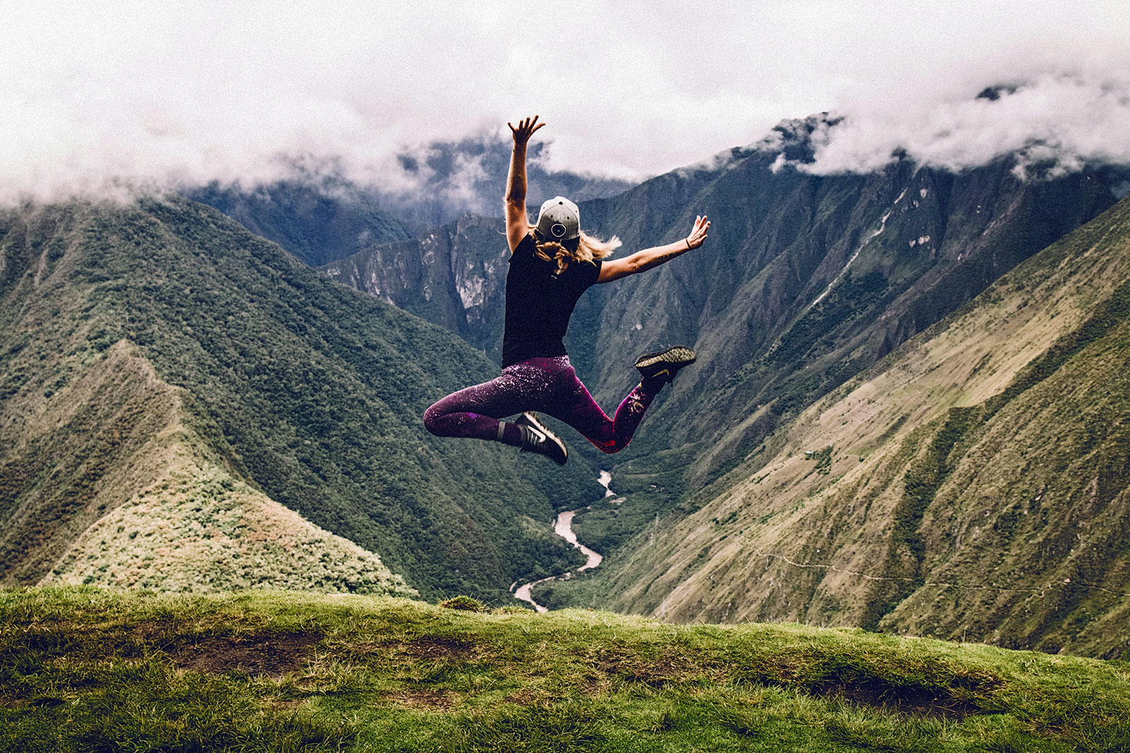 Mulher a saltar, livre, entre duas montanhas, com rio aos pés, vestidas com leggings cor roxa, ténis e t-shirt preta e boné cinzento.